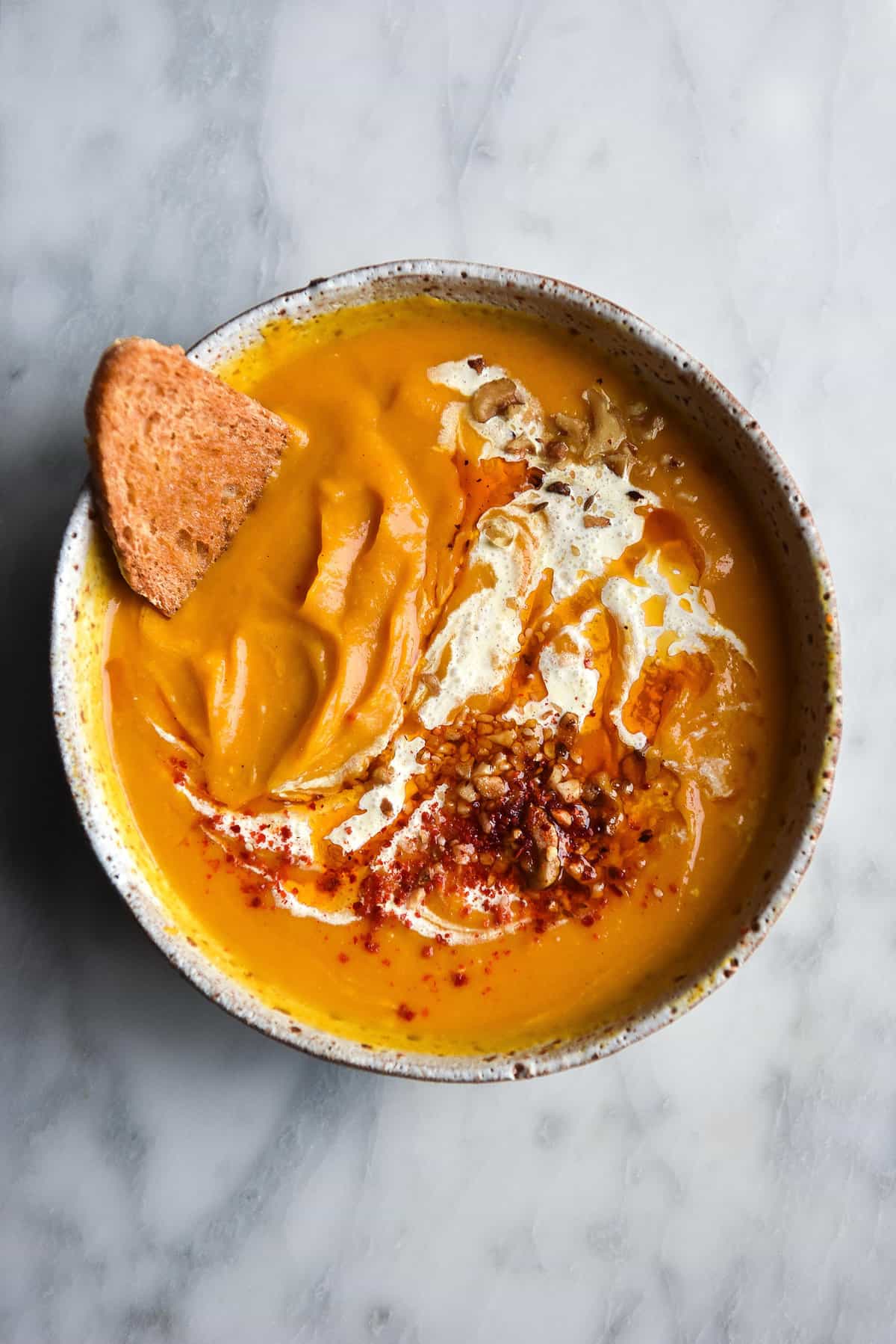 An aerial image of a bowl of pumpkin soup atop a white marble table. The pumpkin soup is topped with swirls of cream and chilli flakes, and a piece of bread dips into the top left corner of the bowl. 