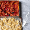 An aerial view of a tray of fresh gluten-free egg pasta and one of cooked tomatoes on a white marble table.