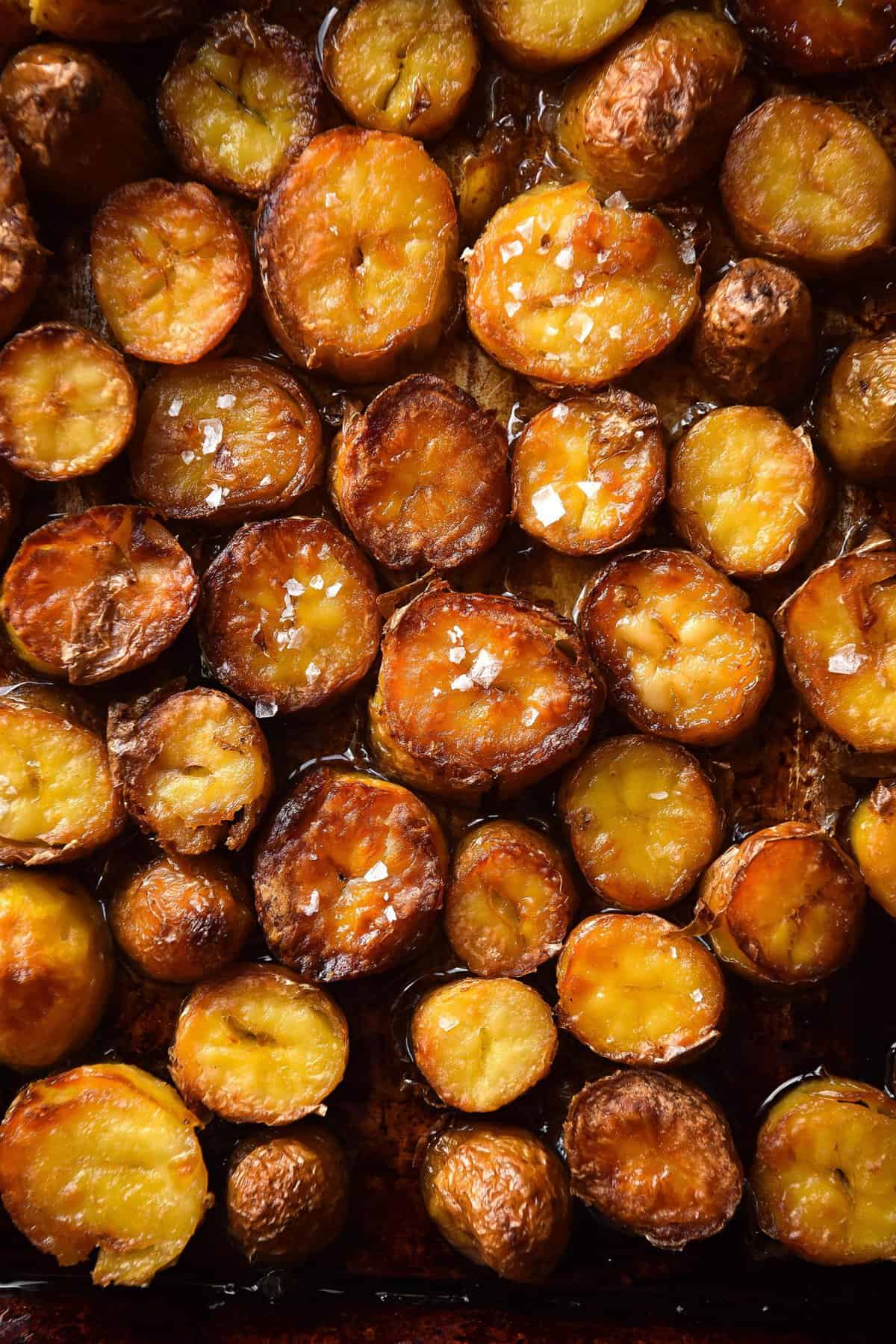An aerial moody image of salt and vinegar roasted potatoes on a black baking tray. The potato slices are golden brown and topped with sea salt flakes