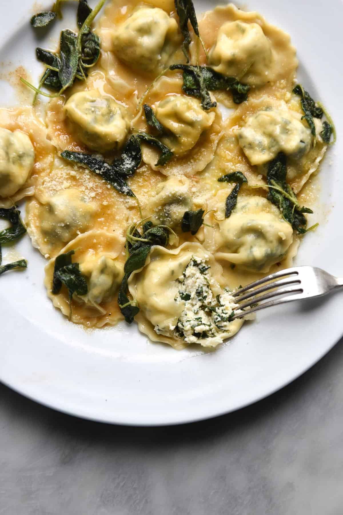 An aerial view of omemade gluten free ravioli with lactose free ricotta and spinach filling and a brown butter crispy sage topping on a white plate. One raviolo is torn open with a fork, revealing the soft ricotta interior.