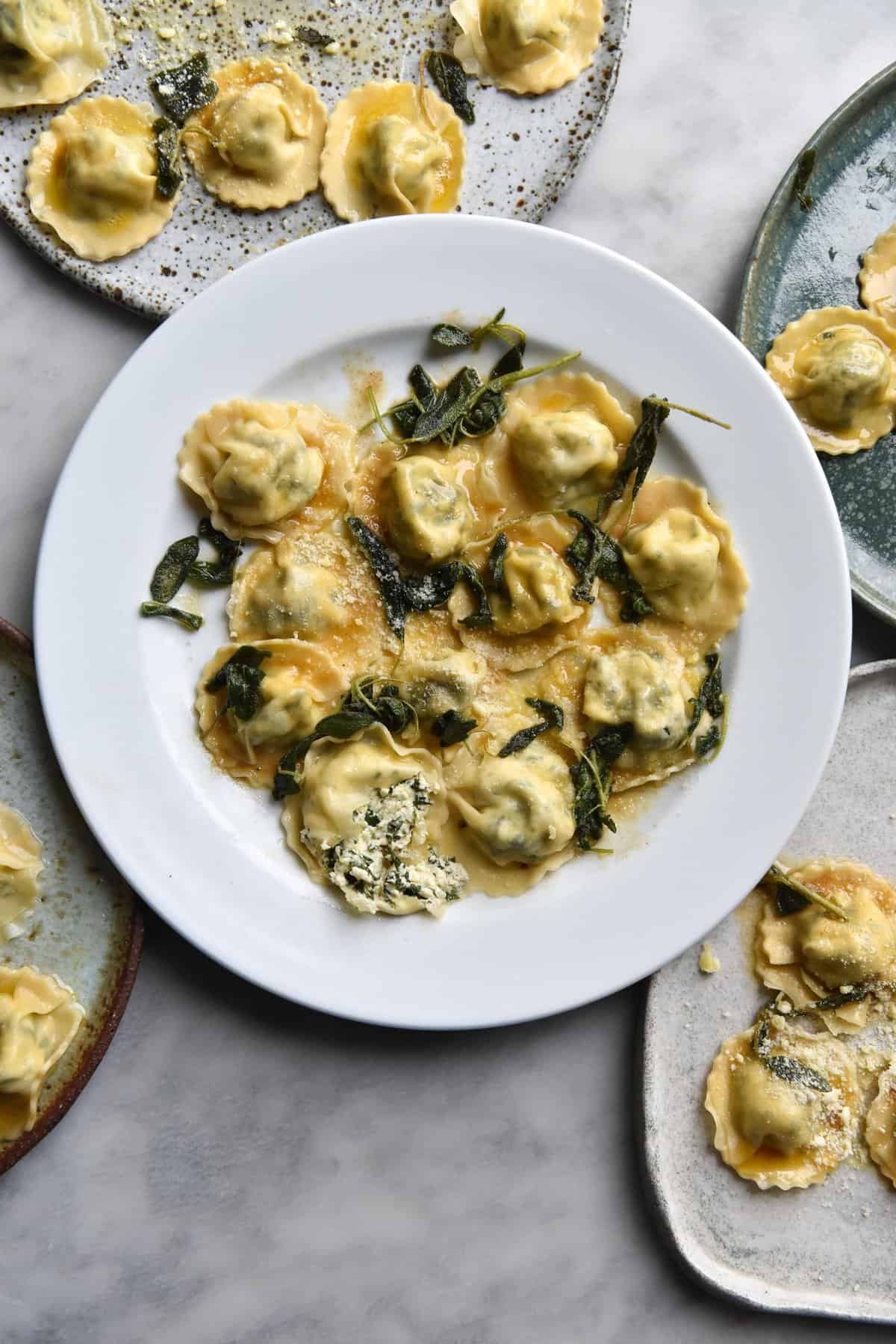 An aerial view of five white ceramic plates topped with gluten free ravioli with a brown butter and sage sauce. The plates sit arranged in a casual pattern around the central plate on a white marble table.