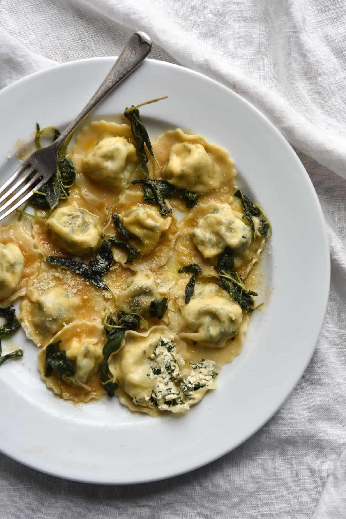 An aerial view of a plate of gluten free ravioli filled with spinach and ricotta and topped with brown butter and sage. The pasta sits in a white dish atop a white linen tablecloth