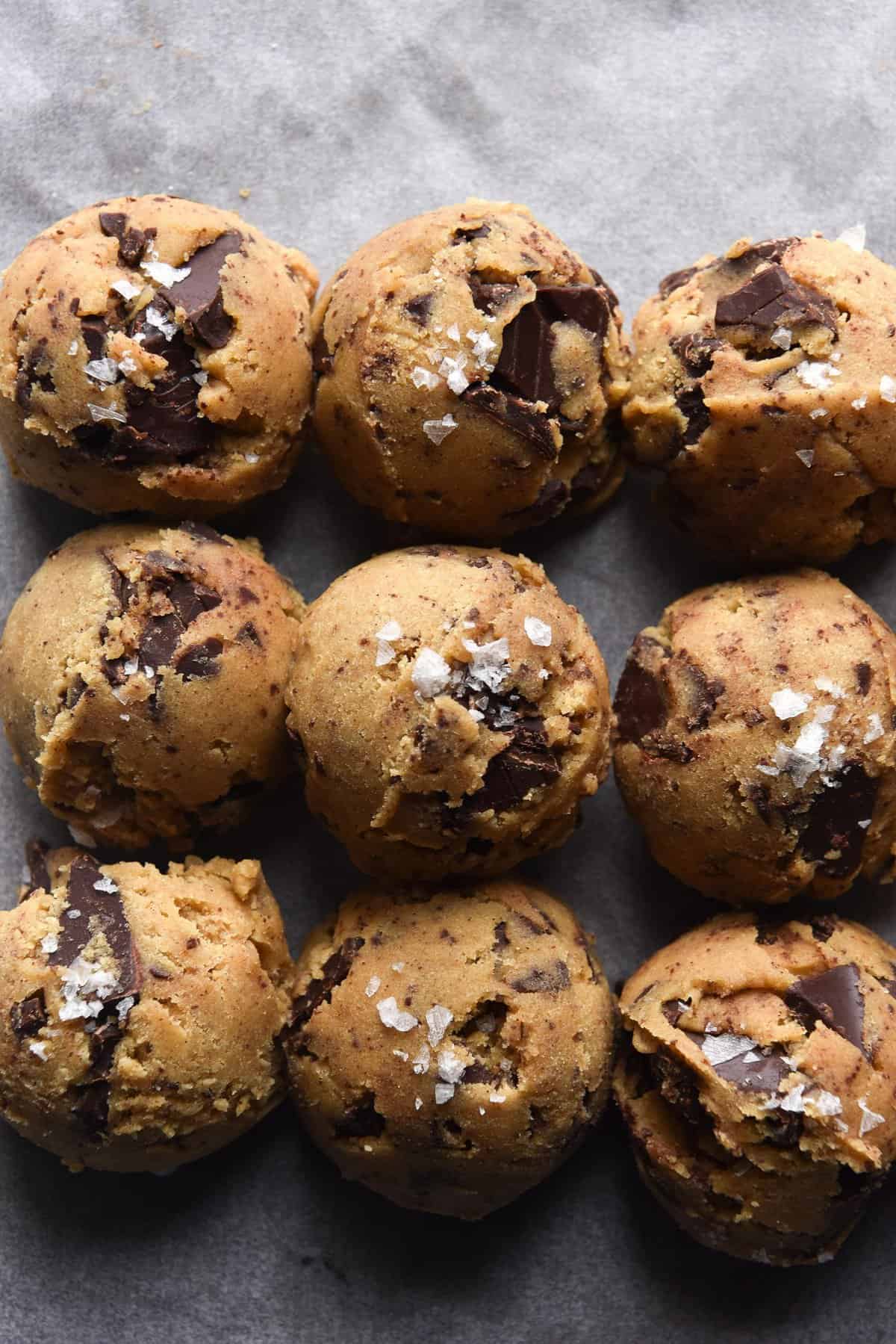 An aerial view of balls of uncooked gluten free choc chip cookie dough on a tray lined with baking paper. Some of the dough balls are sprinkled with salt