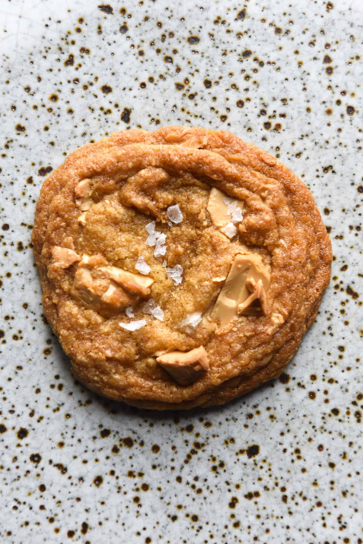 An aerial view of a gluten free choc chip cookie with white chocolate chips and sea salt flakes. The cookie sits atop a white speckled ceramic plate. 
