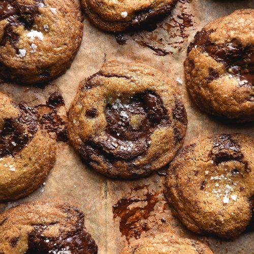 An aerial view of gluten free choc chip cookies without eggs on a sheet of light brown baking paper. The cookies have melted pools of chocolate and sea salt atop each one.