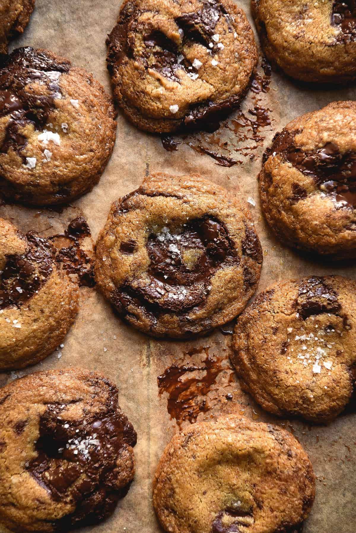 An aerial view of gluten free choc chip cookies without eggs on a sheet of light brown baking paper. The cookies have melted pools of chocolate and sea salt atop each one.