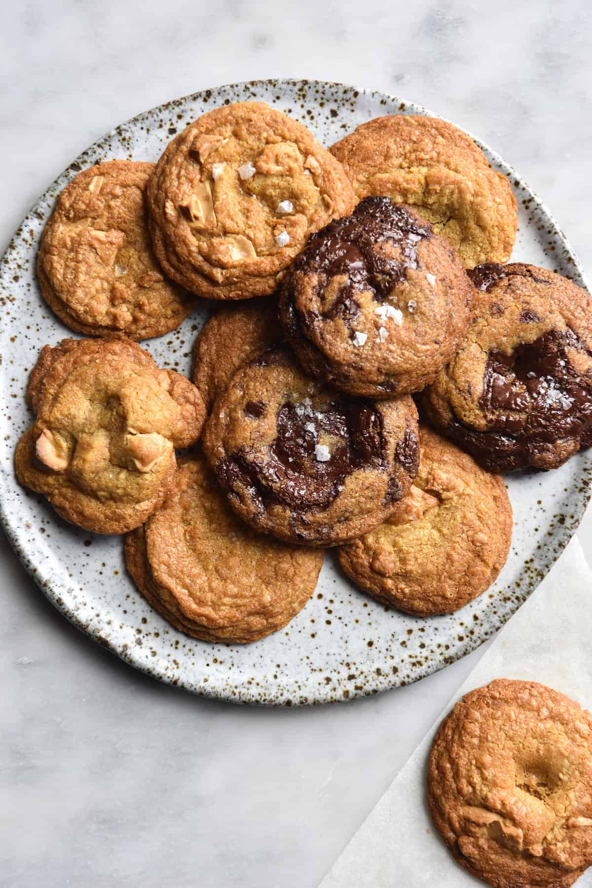 An aerial view of a plate of gluten free choc chip cookies. The cookies sit on a white speckled ceramic plate atop a white marble table. Some of the cookies have melty dark chocolate and some have white chocolate. 