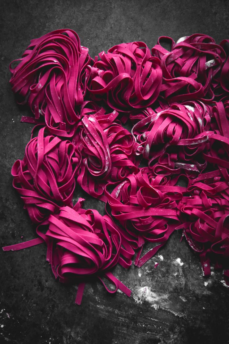 A dark and moody image of freshly rolled gluten-free beetroot pasta atop a dark blue backdrop