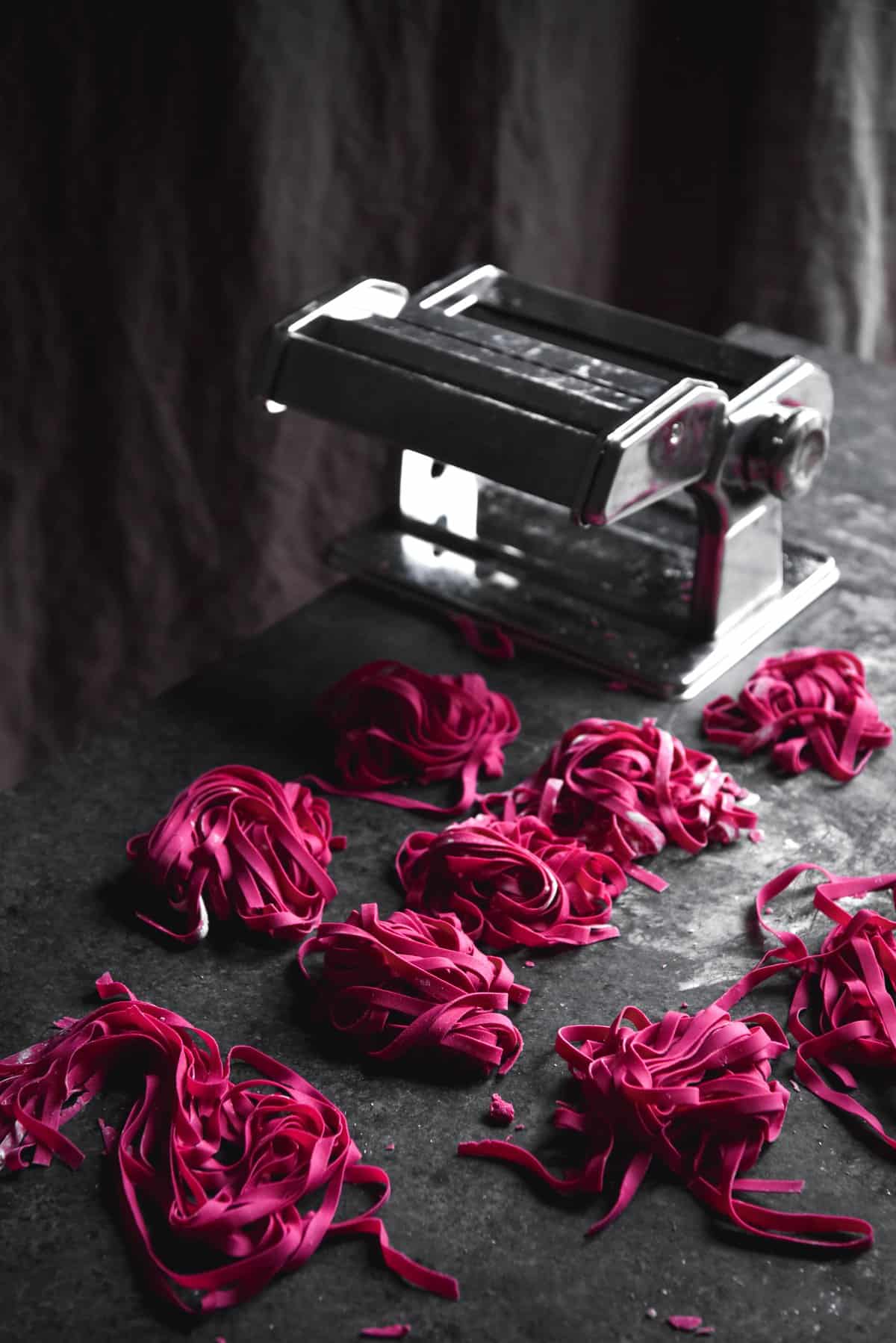 Gluten-free beetroot egg pasta arranged in nests and a pasta machine atop a dark blue backdrop