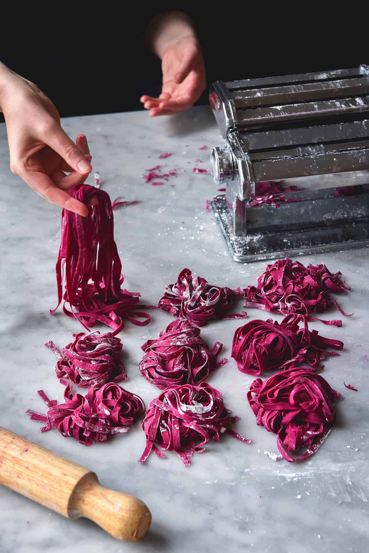 Gluten-free beetroot pasta arranged in nests atop a white marble table. A hand extends from the back corner to hold up the pasta. A pasta machine and a rolling pin surround the nests of pasta.