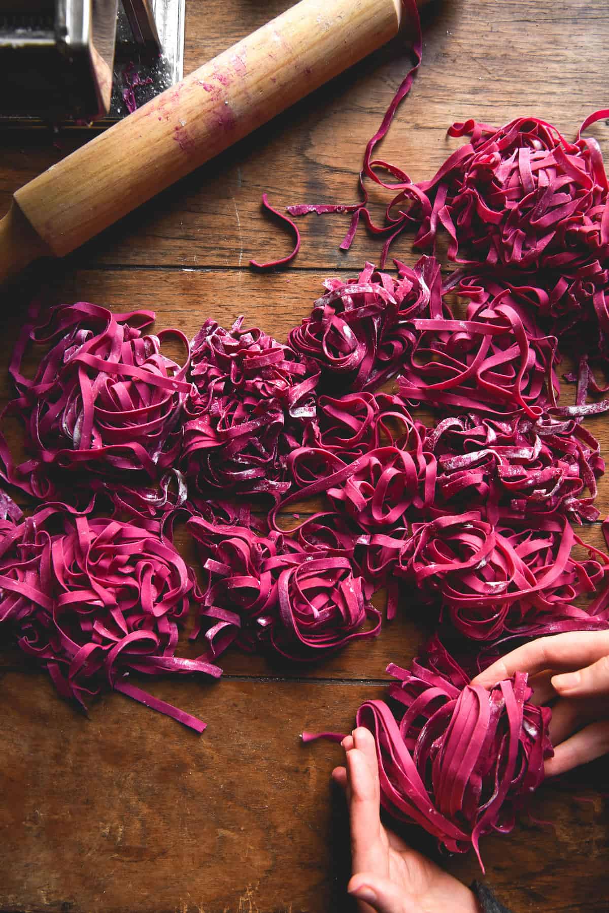 Gluten-free beetroot pasta on a wooden table. A pasta roller and rolling pin sit to the top left of the image.