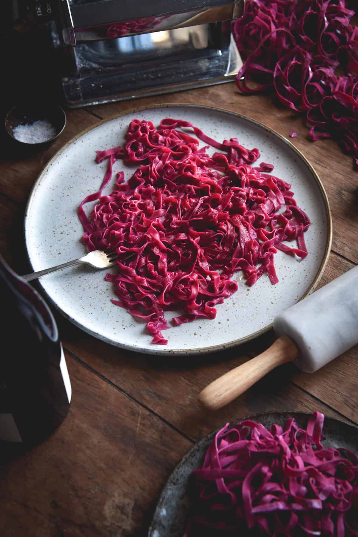 Gluten-free beetroot egg pasta on a white ceramic plate atop a wooden table. Pasta making equipment casually surrounds the plate of pasta.