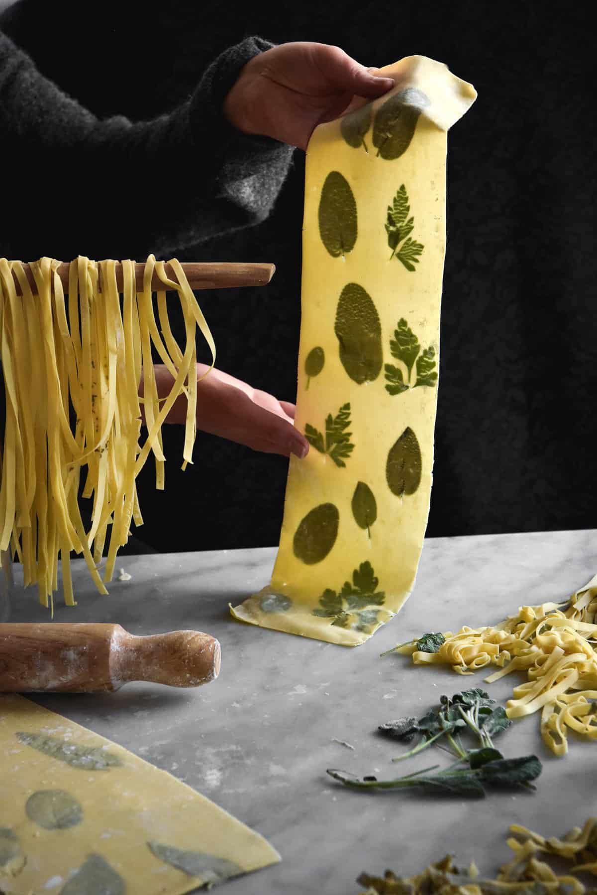 A moody scene of some gluten free egg pasta laminated with sage and parsley leaves. A hand holds the laminated pasta sheet to the right, while fresh fettuccine hands over a spoon to the left. The foreground is strewn with fresh pasta, sage leaves and extra pasta sheets.