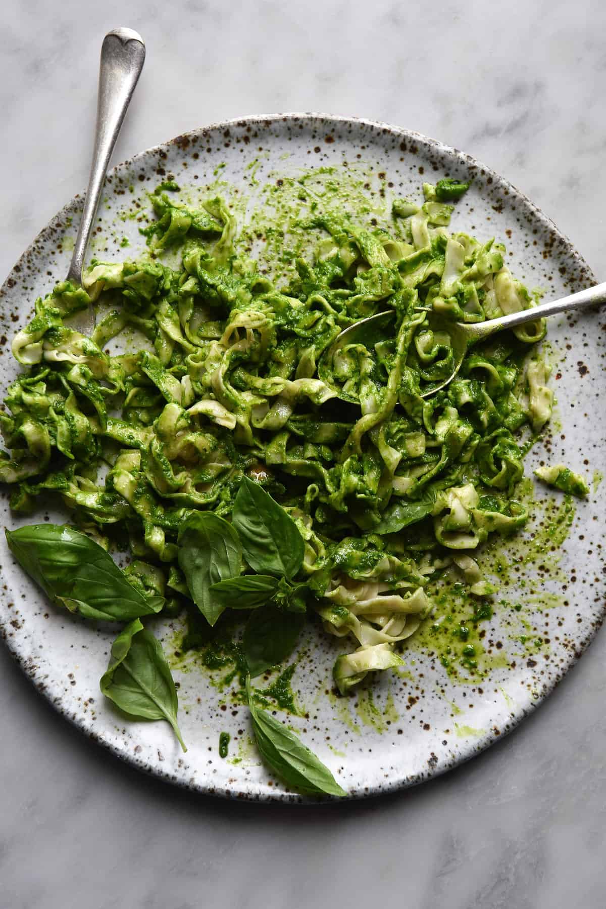 An aerial image of gluten free egg pasta topped with a verdant green kale pesto. The pasta sits casually arranged on a speckled white ceramic plate atop a white marble table