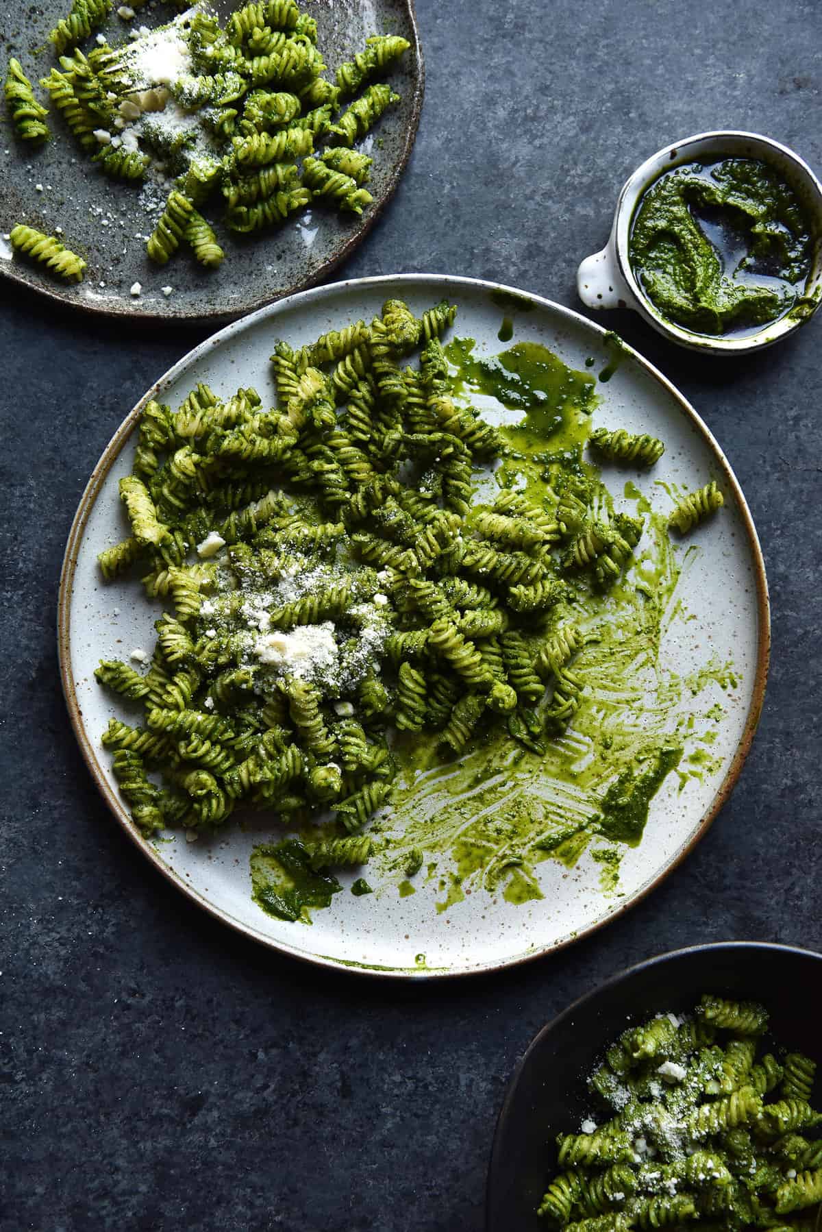 An aerial image image of kale pesto pasta on a white speckled ceramic plate atop a dark blue backdrop