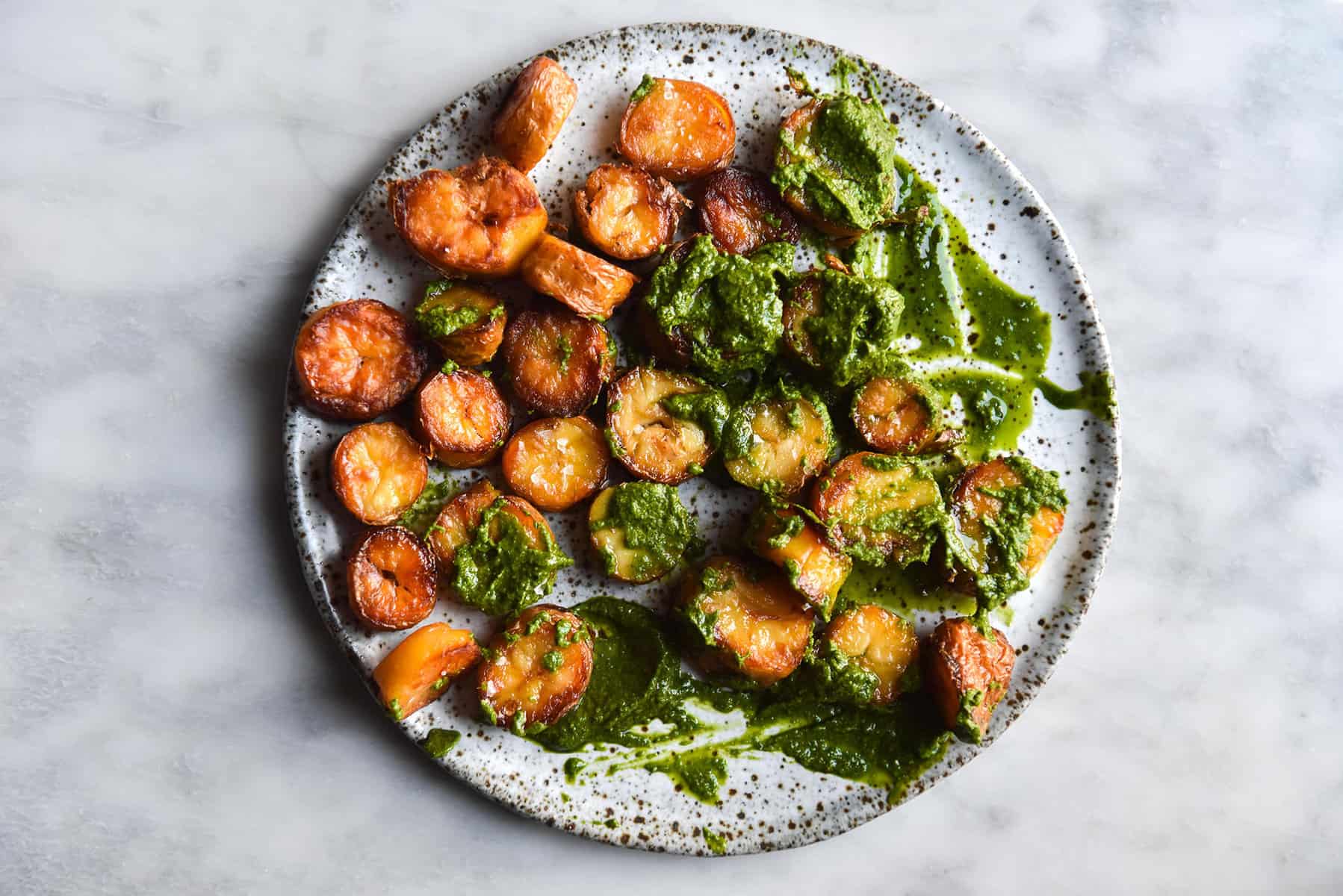 An aerial image of crispy roasted potatoes smothered in kale pesto on a white speckled ceramic plate atop a white marble table. 