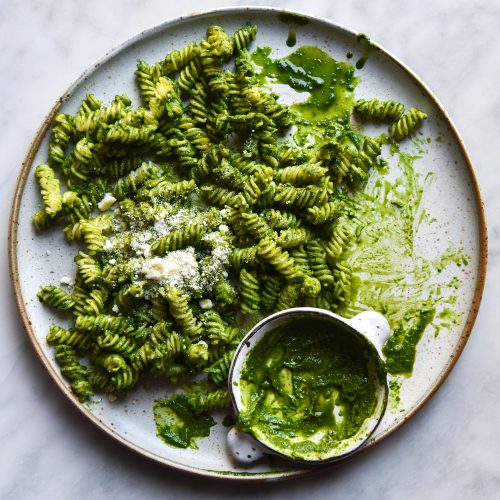 An aerial close up view of a white ceramic plate topped with verdant green kale pesto pasta. A small bowl of kale pesto sits to the bottom of the plate, which sits atop a white marble table