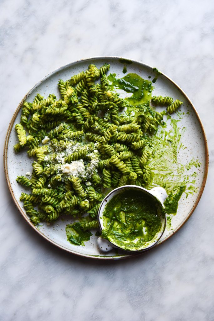 An aerial close up view of a white ceramic plate topped with verdant green kale pesto pasta. A small bowl of kale pesto sits to the bottom of the plate, which sits atop a white marble table
