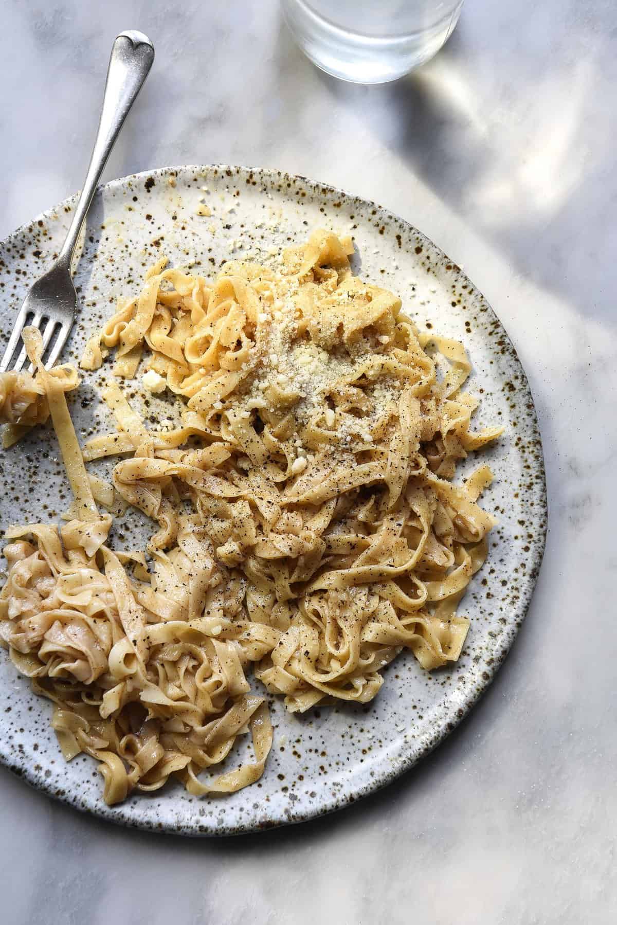 An aerial view of a white speckled ceramic plate on a white marble table topped with gluten free egg pasta. The pasta is topped with a cacio e pepe sauce. 