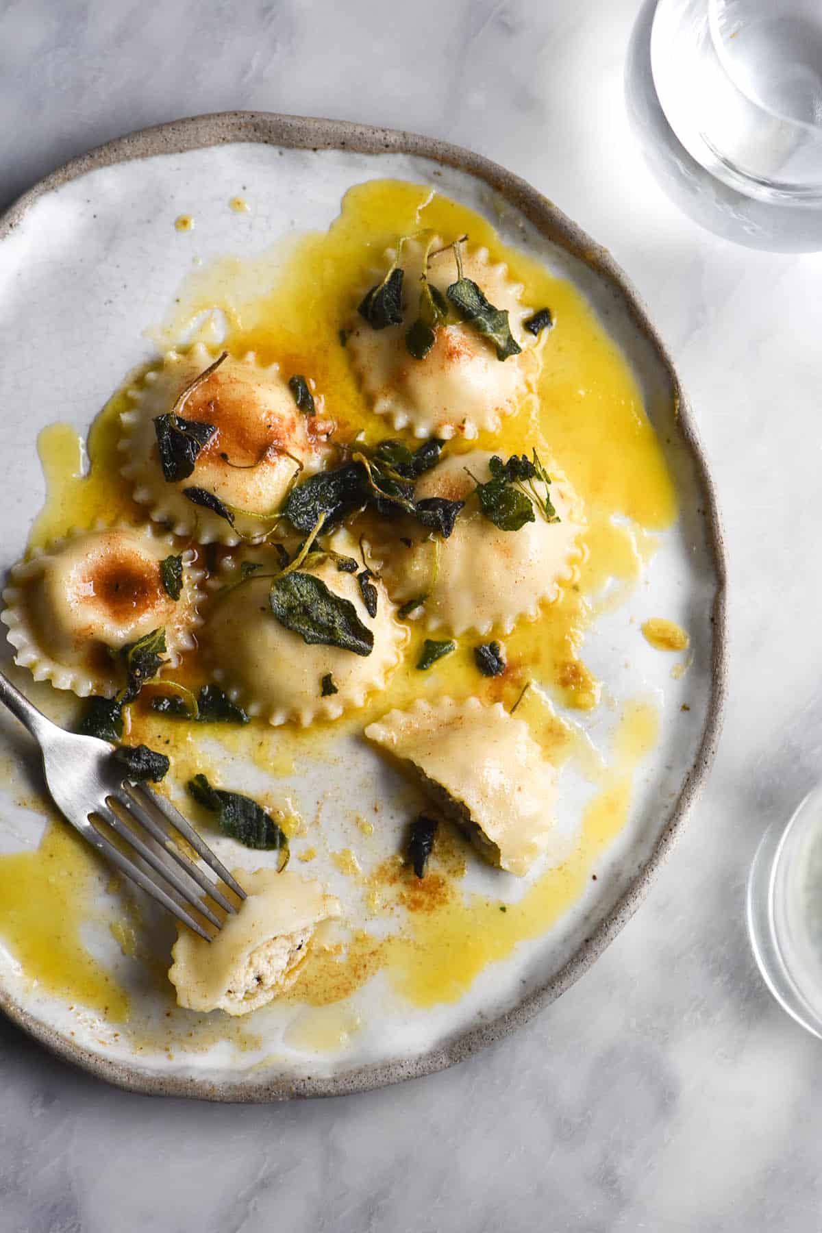 An aerial view of a white ceramic plate topped with gluten free spinach and ricotta stuffed ravioli and topped with crispy sage and brown butter. The plate sits on a white marble table and is surrounded by water glasses