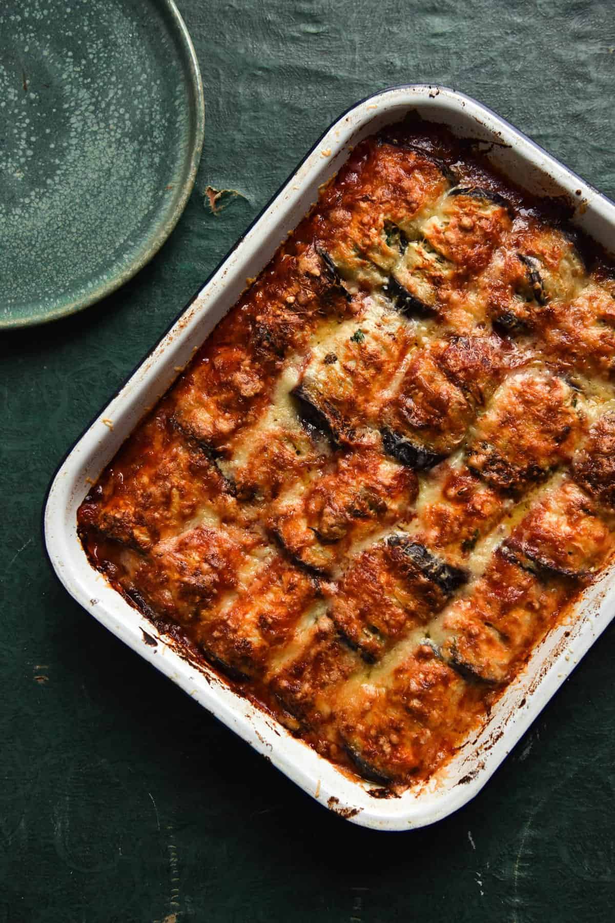 A tray of vegan sausage stuffed involtini in a melty cheesy bake atop an olive green table. A green plate sits to the top left of the image.