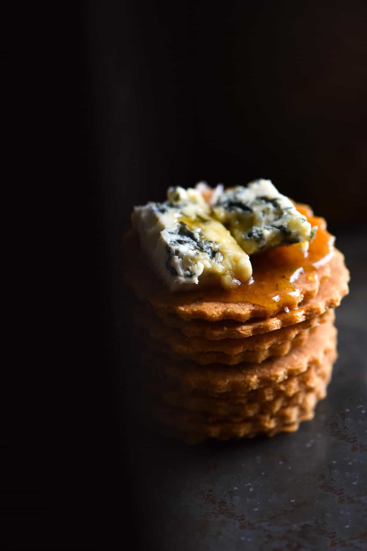 A dark and moody image of a stack of gluten free almond crackers. The top cracker is finished with a lump of blue cheese and a drizzle of honey. 