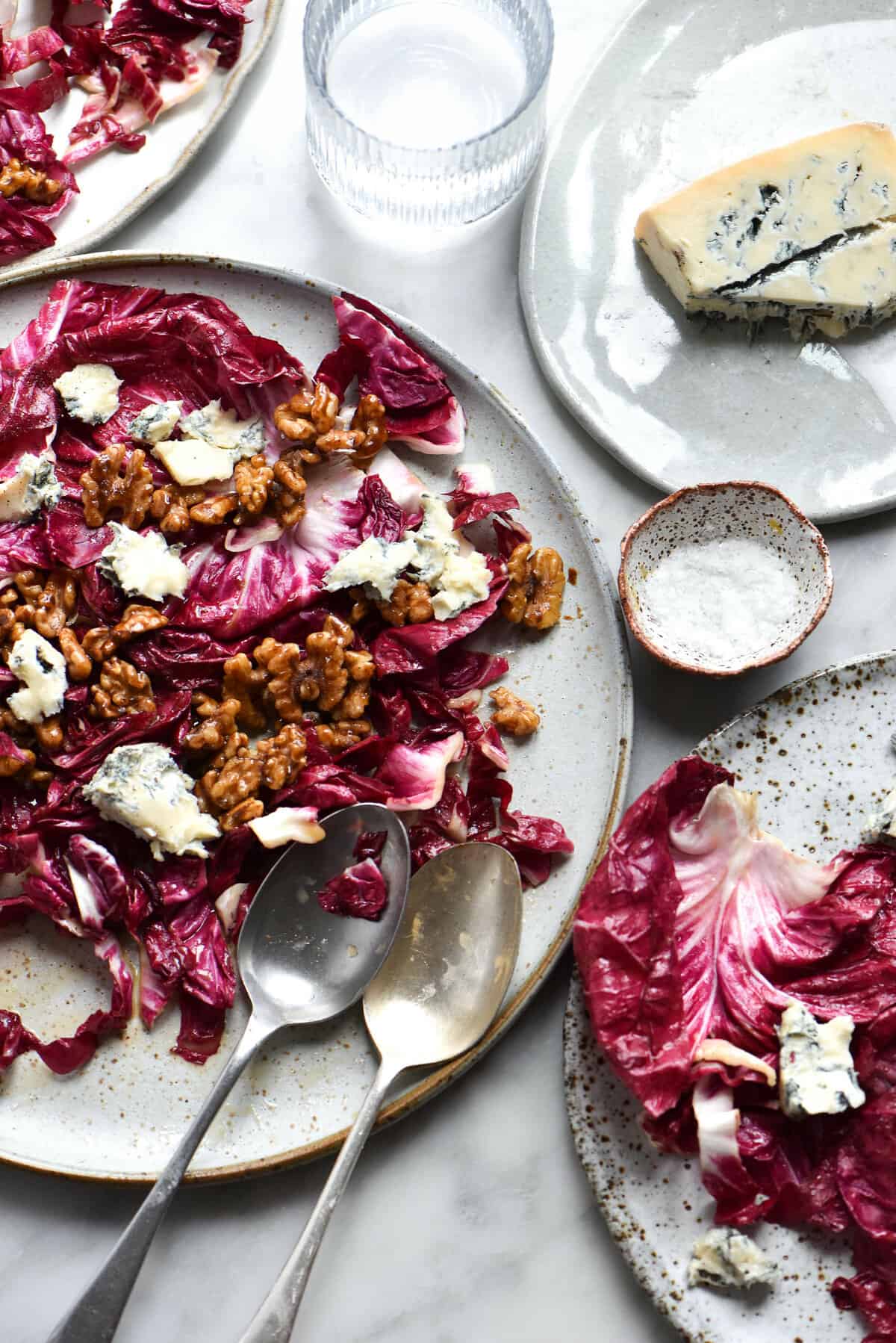 Radicchio salad with blue cheese, honey cinnamon walnuts and a sherry vinaigrette against a white marble backdrop. The salad is plated on a white ceramic plate and other plates of salad surround the main one. A hunk of blue cheese sits to the top right of the image and water glasses cast a soft light across the top of the image