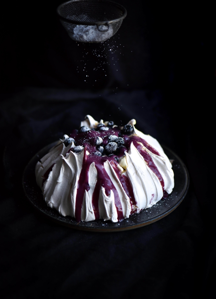 A tall pavlova topped with lactose free blueberry curd and a sprinkle of icing sugar against a black backdrop
