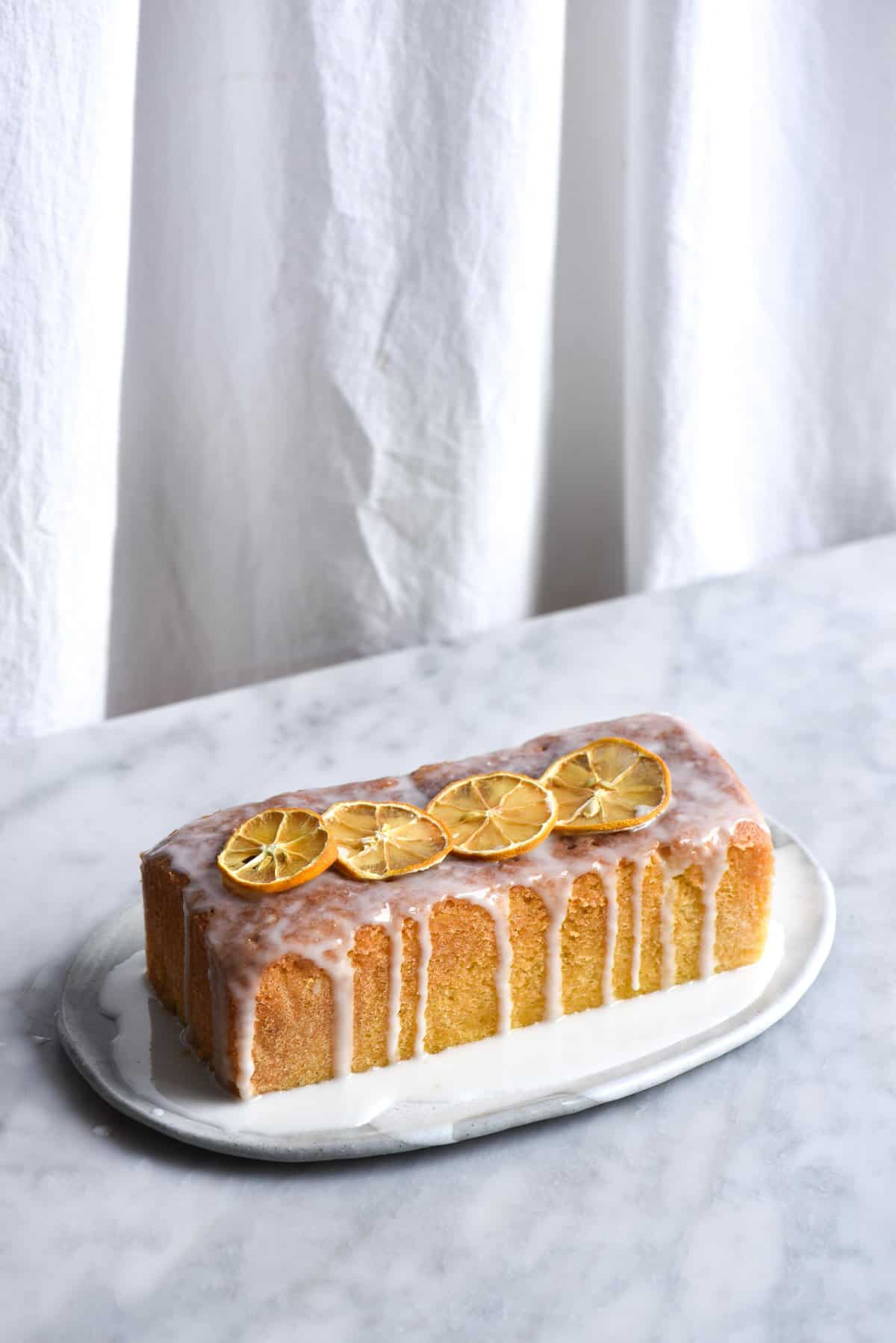 Gluten free lemon drizzle cake loaf with dripping lemon icing against a white marble backdrop