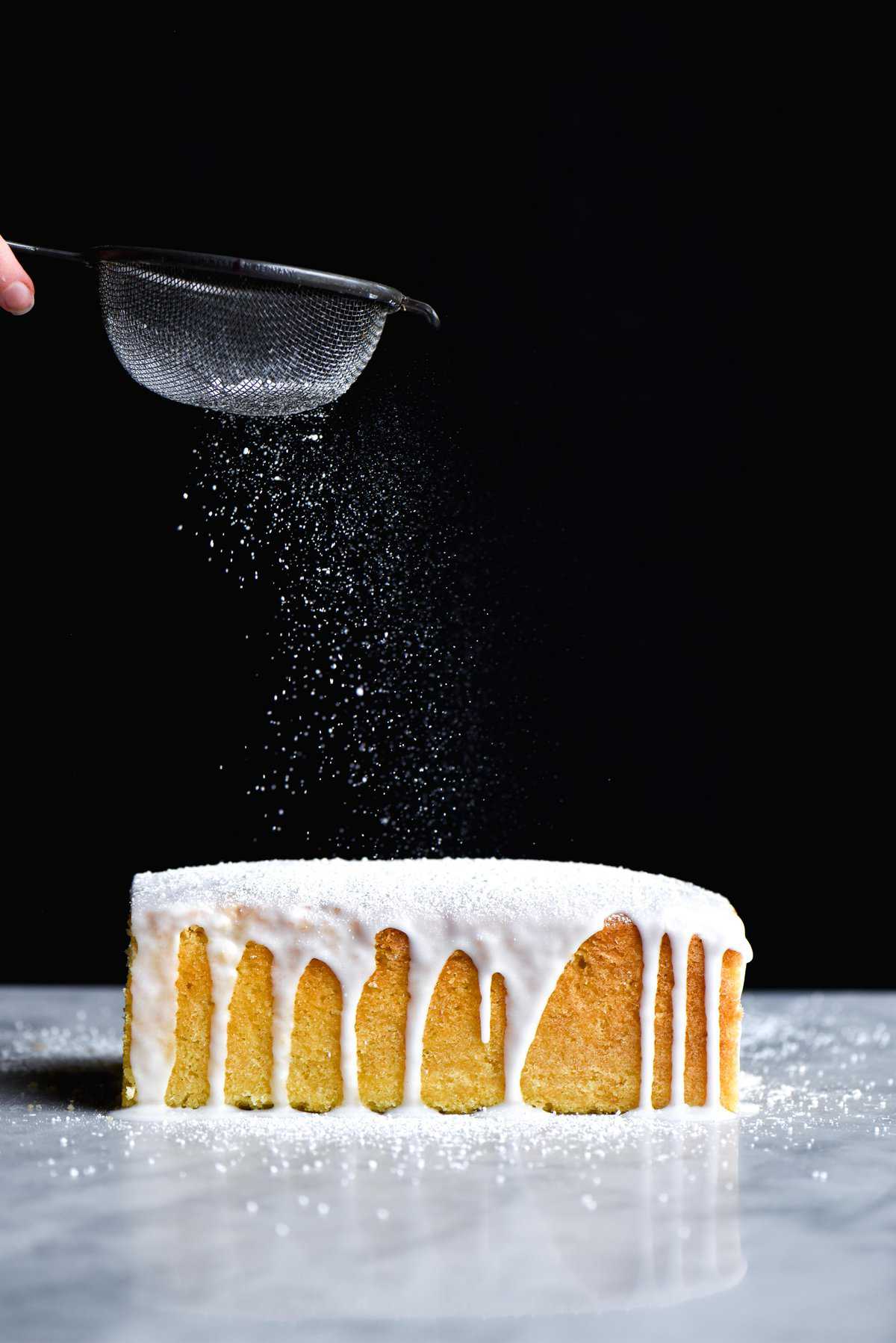 A side on view of a gluten free drizzle cake being sprinkled with icing sugar against a dark backdrop