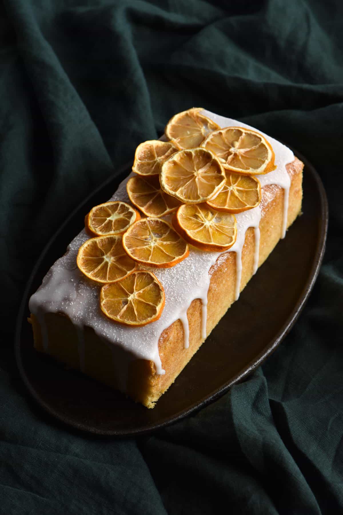 A side on view of a lemon drizzle cake topped with dehydrated lemon slices against a forest green linen backdrop