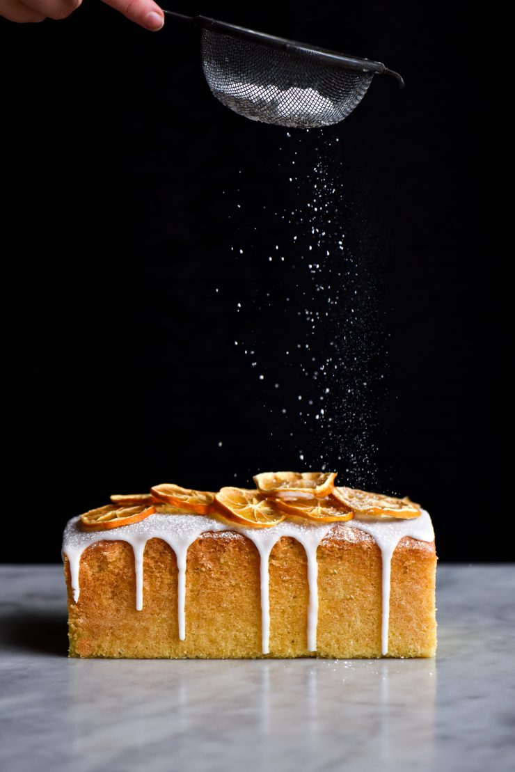 A side on view of a lemon drizzle cake topped with dehydrated lemons and lemon drizzle icing, being dusted with icing sugar against a dark background