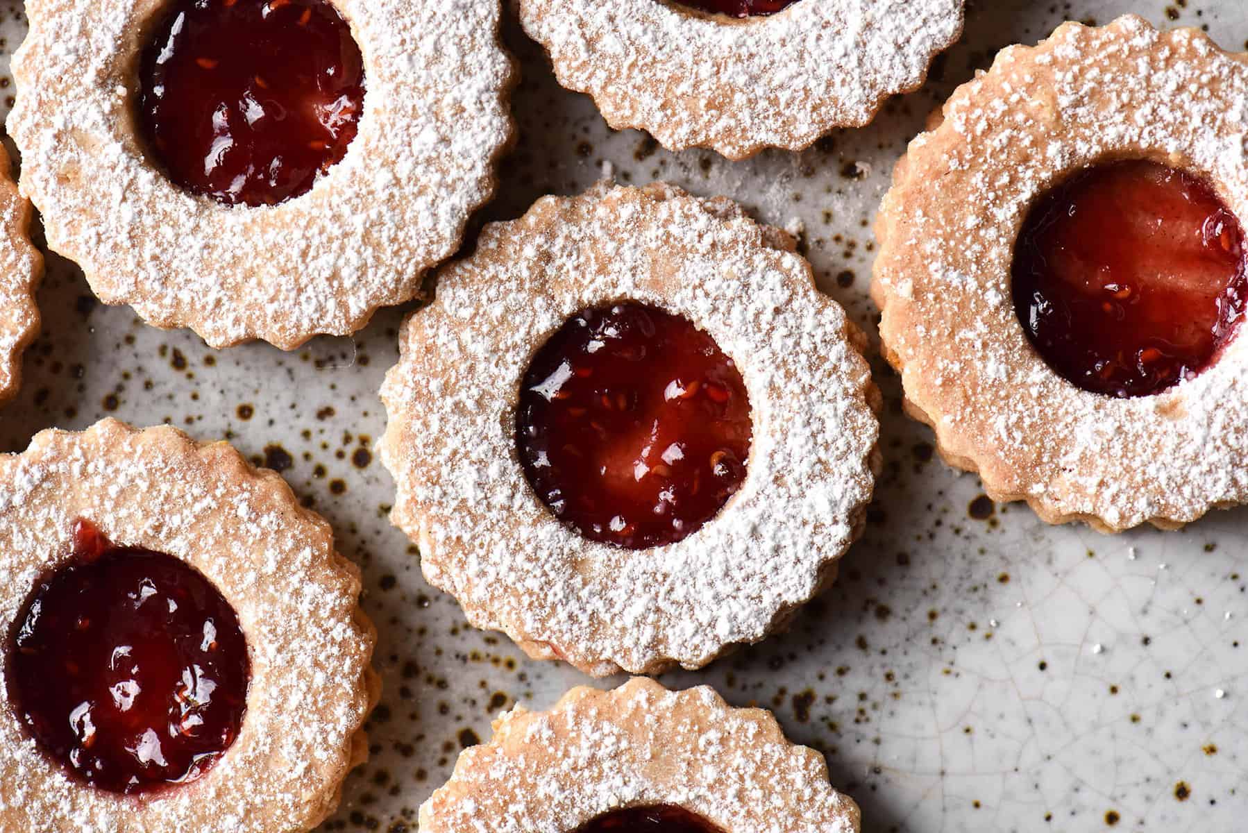 Gluten free linzer cookies against a white ceramic backdrop. The cookies are filled with raspberry jam and sprinkled with icing sugar. 