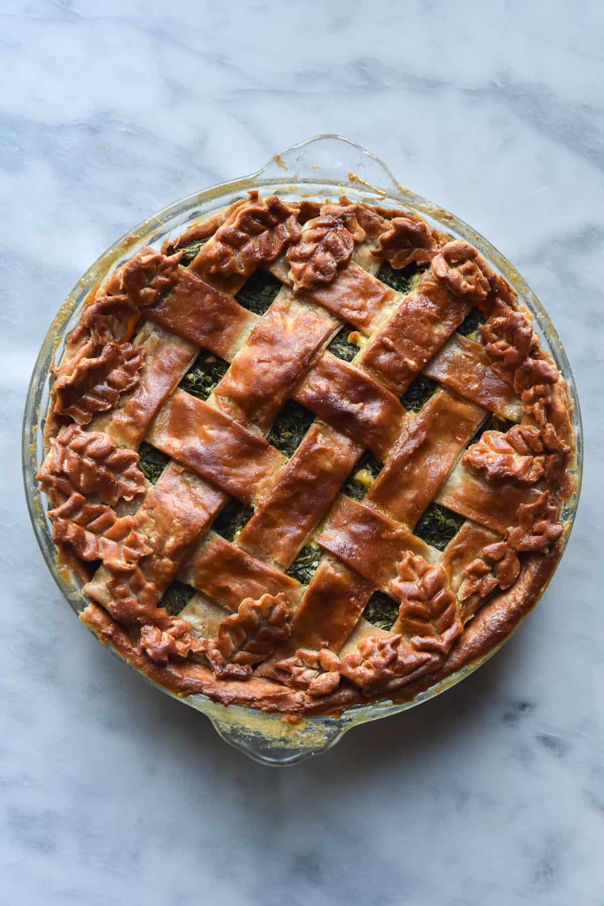 A gluten free greens pie with latticed flaky pastry against a white marble backdrop