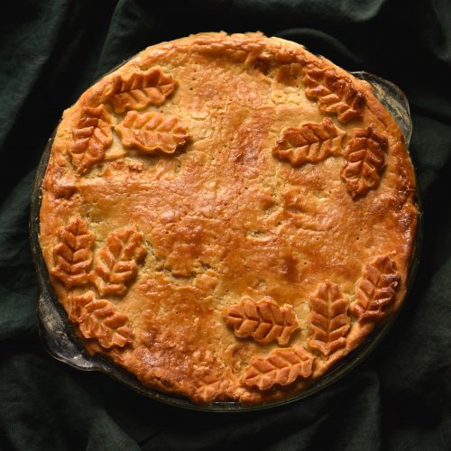 A close up of a gluten free pie with flaky pastry and ornamental pastry leaves against a green linen backdrop