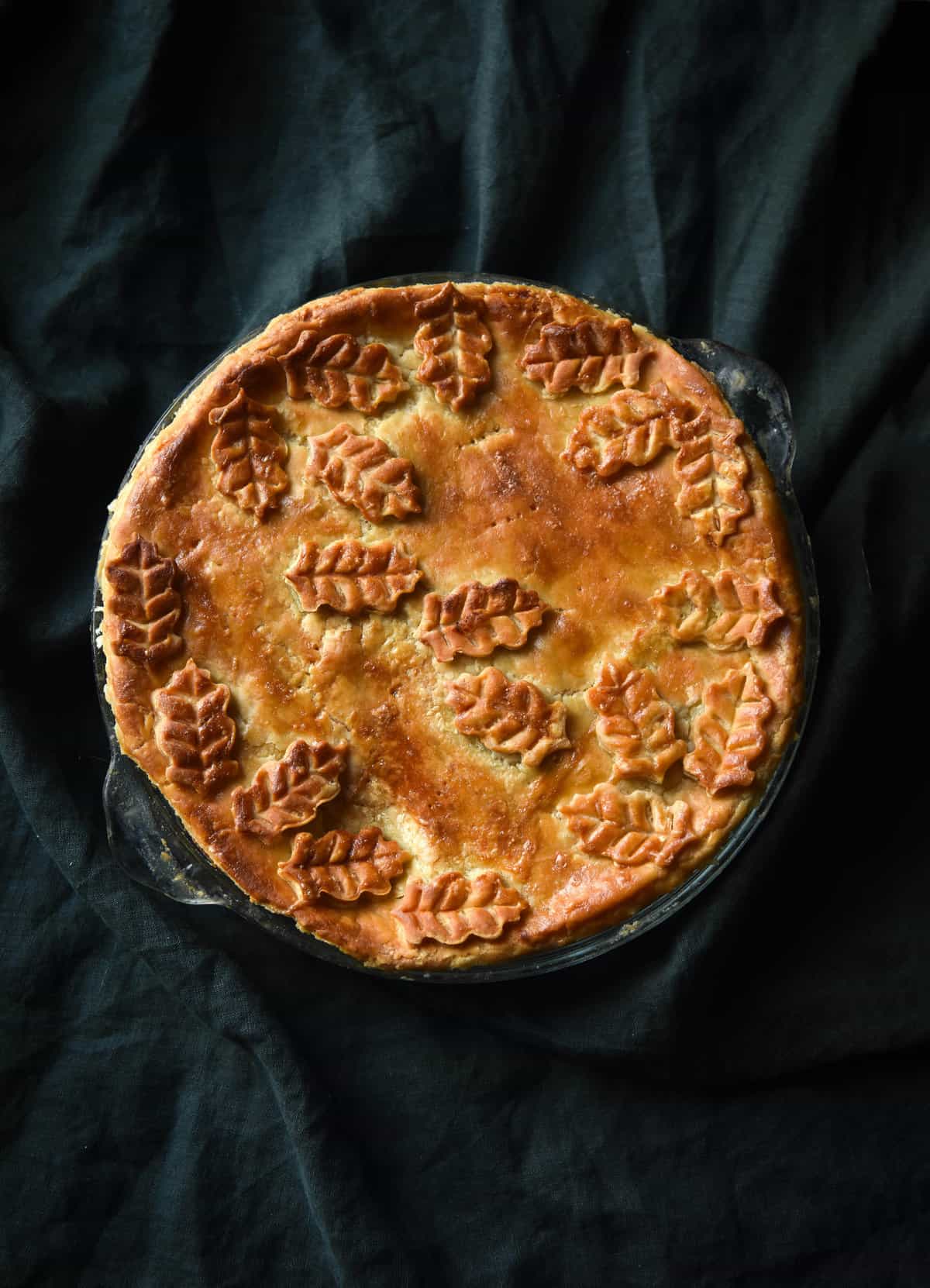 A gluten free greens and feta pie with flaky pastry, adorned with pastry leaves and styled against a forrest green backdrop