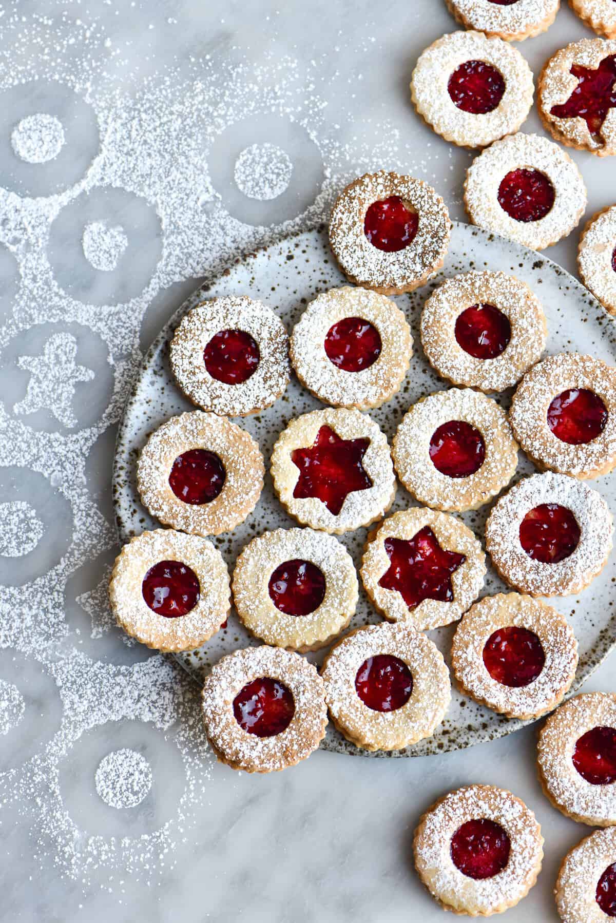 Gluten free linzer cookies with raspberry jam, sprinkled with icing sugar and set against an icing sugar sprinkled white marble backdrop