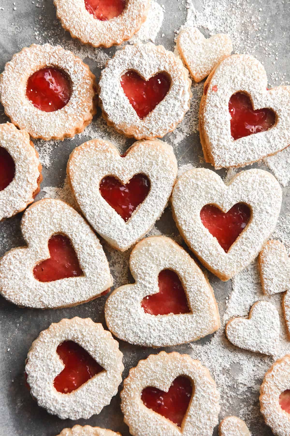 A light and airy image of a tray of gluten free linzer cookies filled with raspberry jam. The cookies are cut in heart shapes with a small heart cut out of the centre, exposing the jam filling. The top are dusted with icing sugar. 