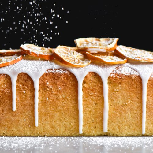 A side on image of a gluten free lemon drizzle cake being sprinkled with icing sugar. The cake sits atop a white marble table against a black backdrop and is topped with drizzled icing and dried lemon slices. Icing sugar sprinkles down from the top of the image