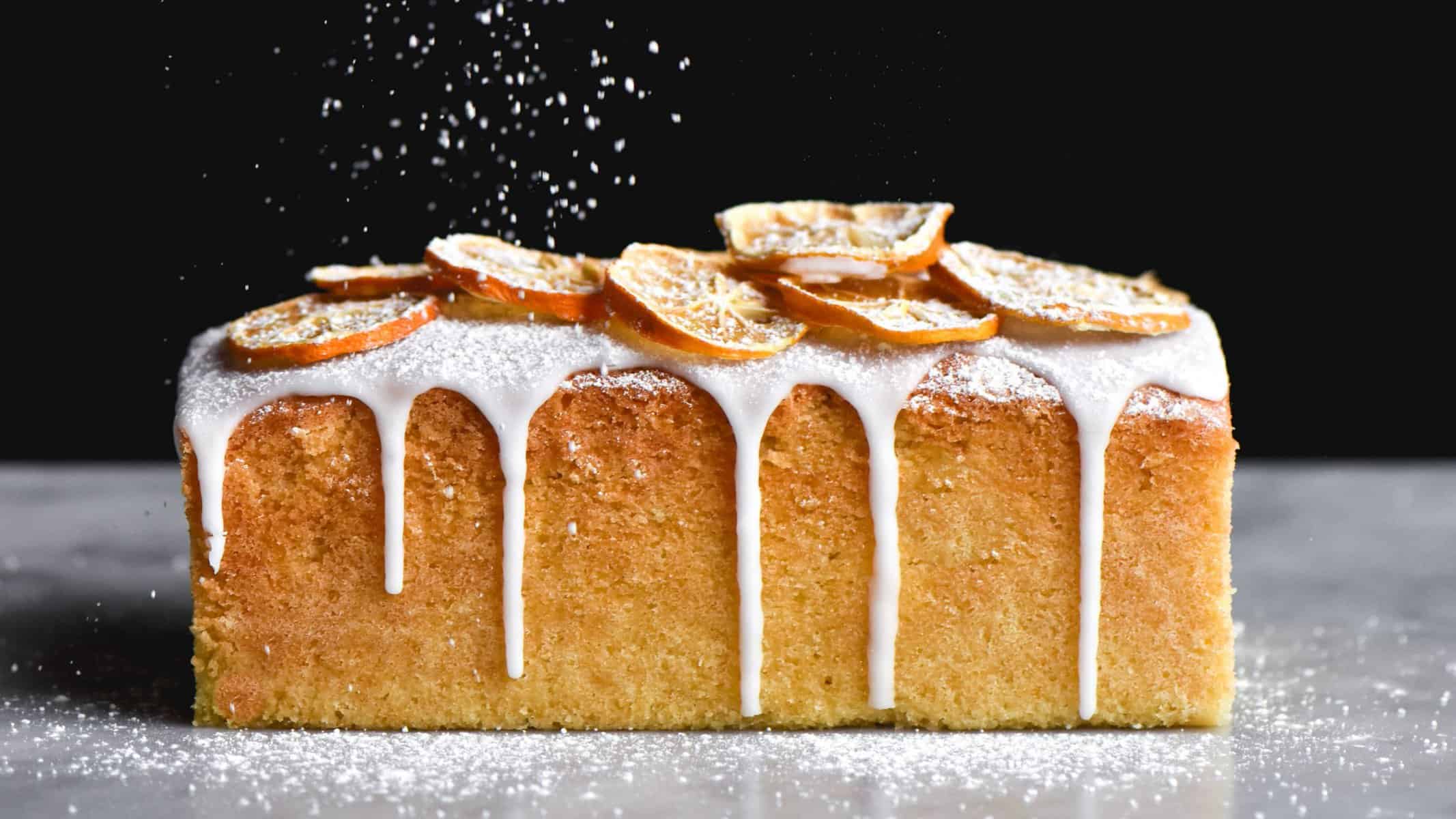 A side on image of a gluten free lemon drizzle cake being sprinkled with icing sugar. The cake sits atop a white marble table against a black backdrop and is topped with drizzled icing and dried lemon slices. Icing sugar sprinkles down from the top of the image