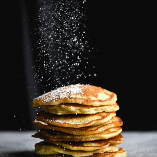 A side on view of gluten free sourdough pancakes being sprinkled with icing sugar against a black backdrop