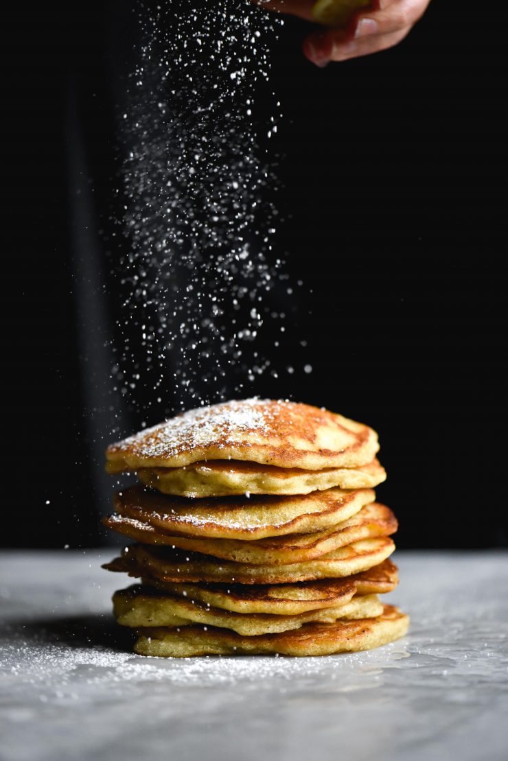 A side on view of gluten free sourdough pancakes being sprinkled with icing sugar against a black backdrop