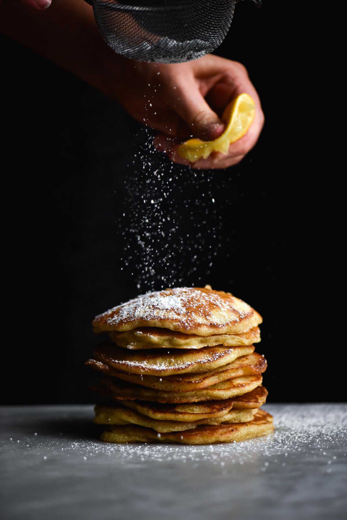 A side on view of a stack of gluten free sourdough pancakes being sprinkled with lemon juice and icing sugar against a dark backdrop
