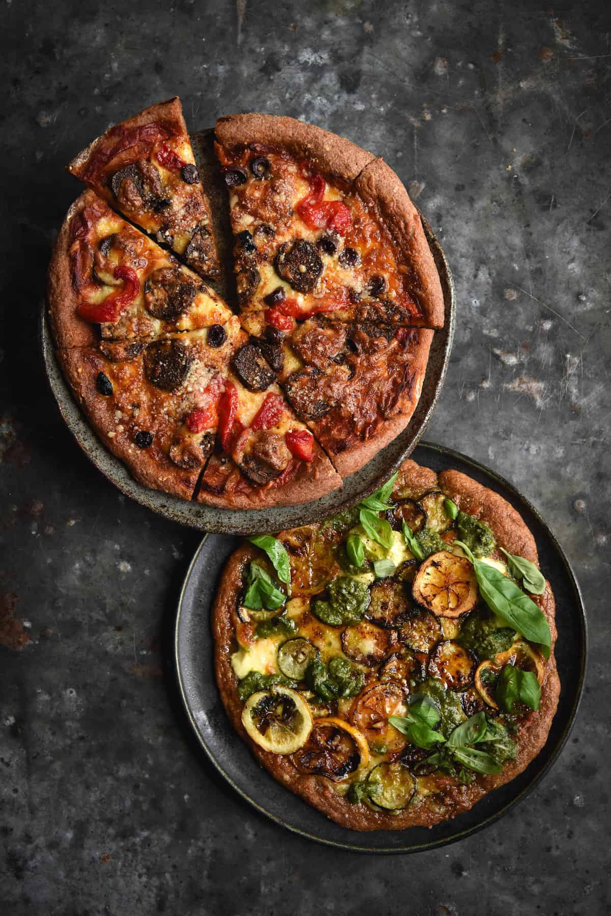 Two gluten free sourdough pizza bases against a blue mottled backdrop. One is a red pizza topped with vegetarian sausage, roasted capsicums and cheese, the other is a green pizza, topped with char grilled zucchini and lemons, pesto and basil