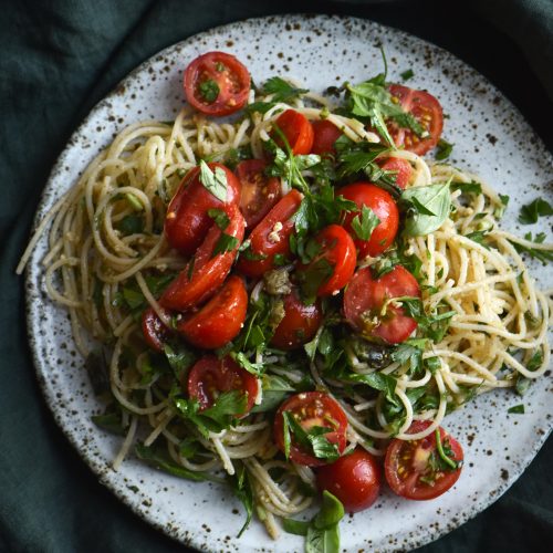 FODMAP friendly pasta with a brown butter, caper, chilli, lemon and herb sauce topped with fresh summer tomatoes, served on a white ceramic plate against a green linen table cloth