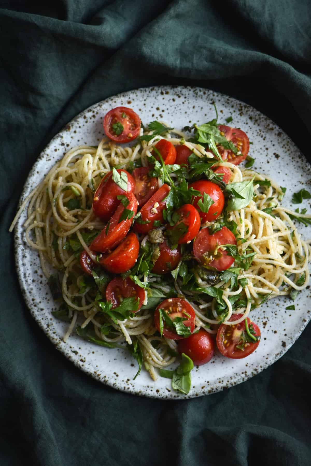 FODMAP friendly pasta with a brown butter, caper, chilli, lemon and herb sauce topped with fresh summer tomatoes, served on a white ceramic plate against a green linen table cloth