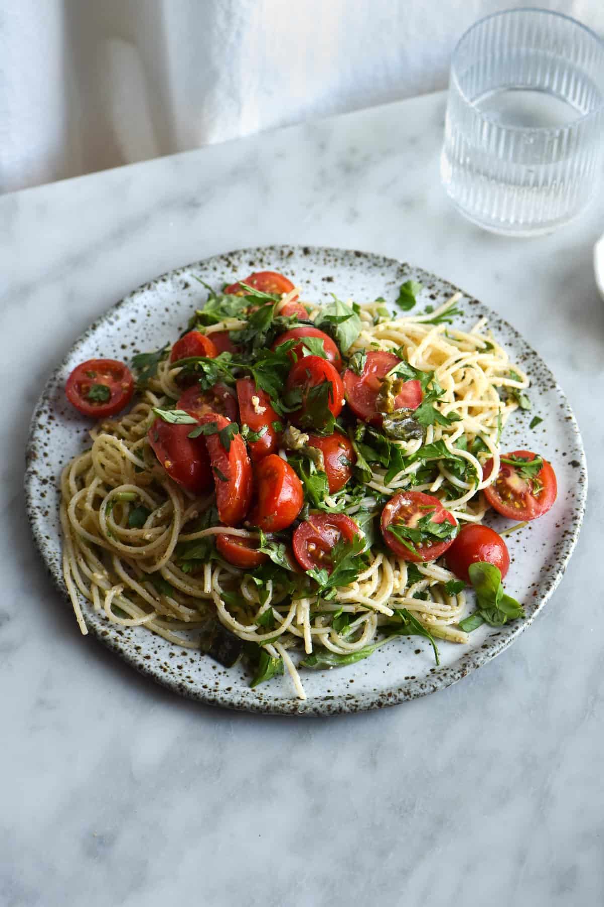 A plate of gluten free pasta with brown butter, lemon and herb sauce, topped with fresh summer tomatoes. Set against a white marble backdrop on a white ceramic plate.