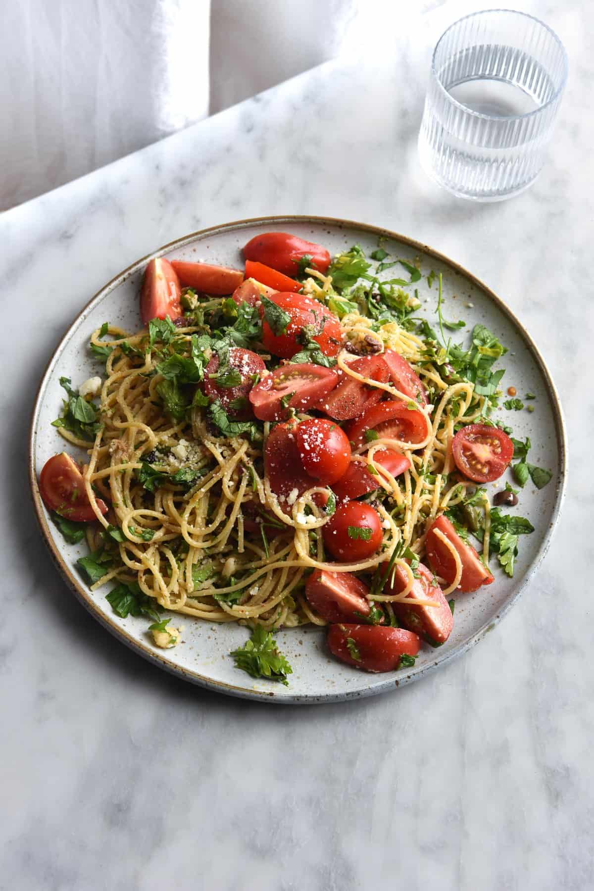A plate of gluten free pasta with a brown butter, lemon, chilli and herb sauce, topped with fresh summer tomatoes against a white marble backdrop