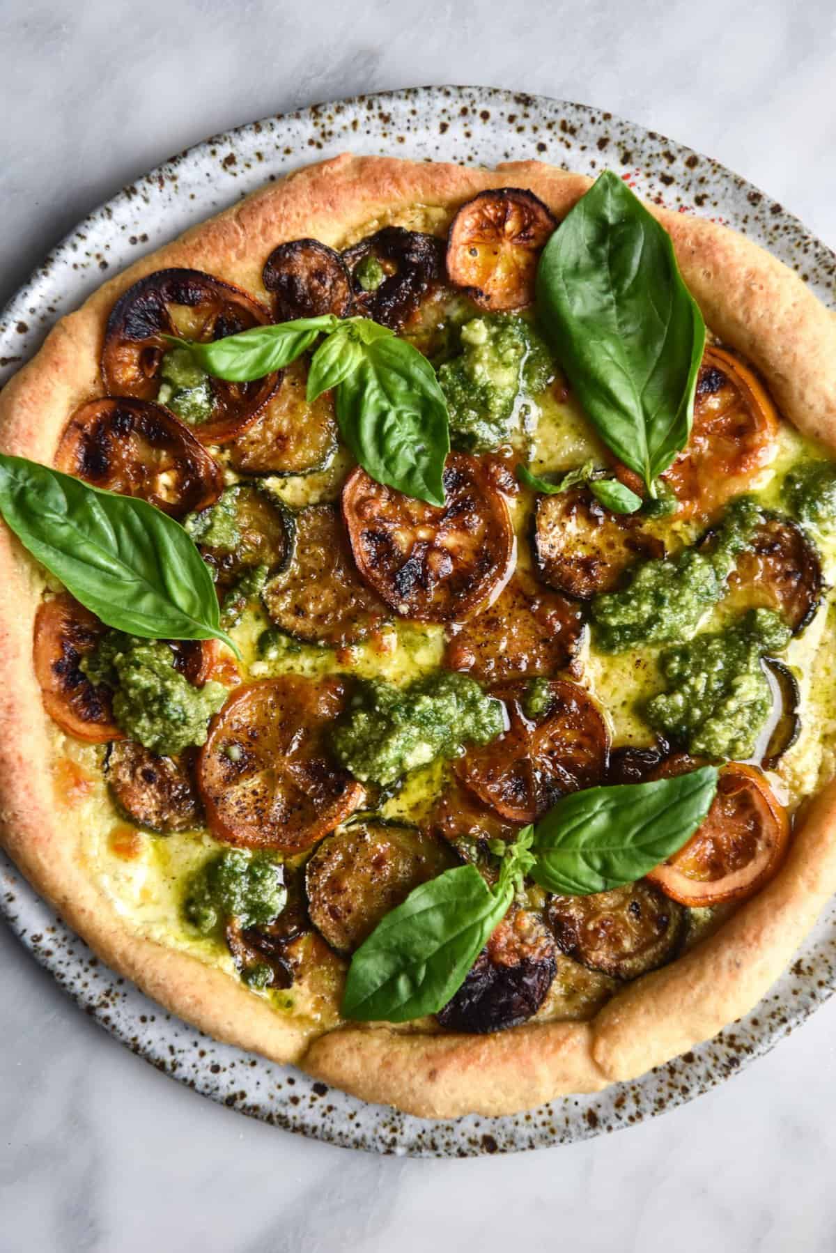 An aerial view of a FODMAP friendly, gluten free sourdough pizza topped with caramelised lemons, zucchini coins, FODMAP friendly pesto, melted cheese and extra basil. The pizza sits on a speckled white plate against a white marble backdrop