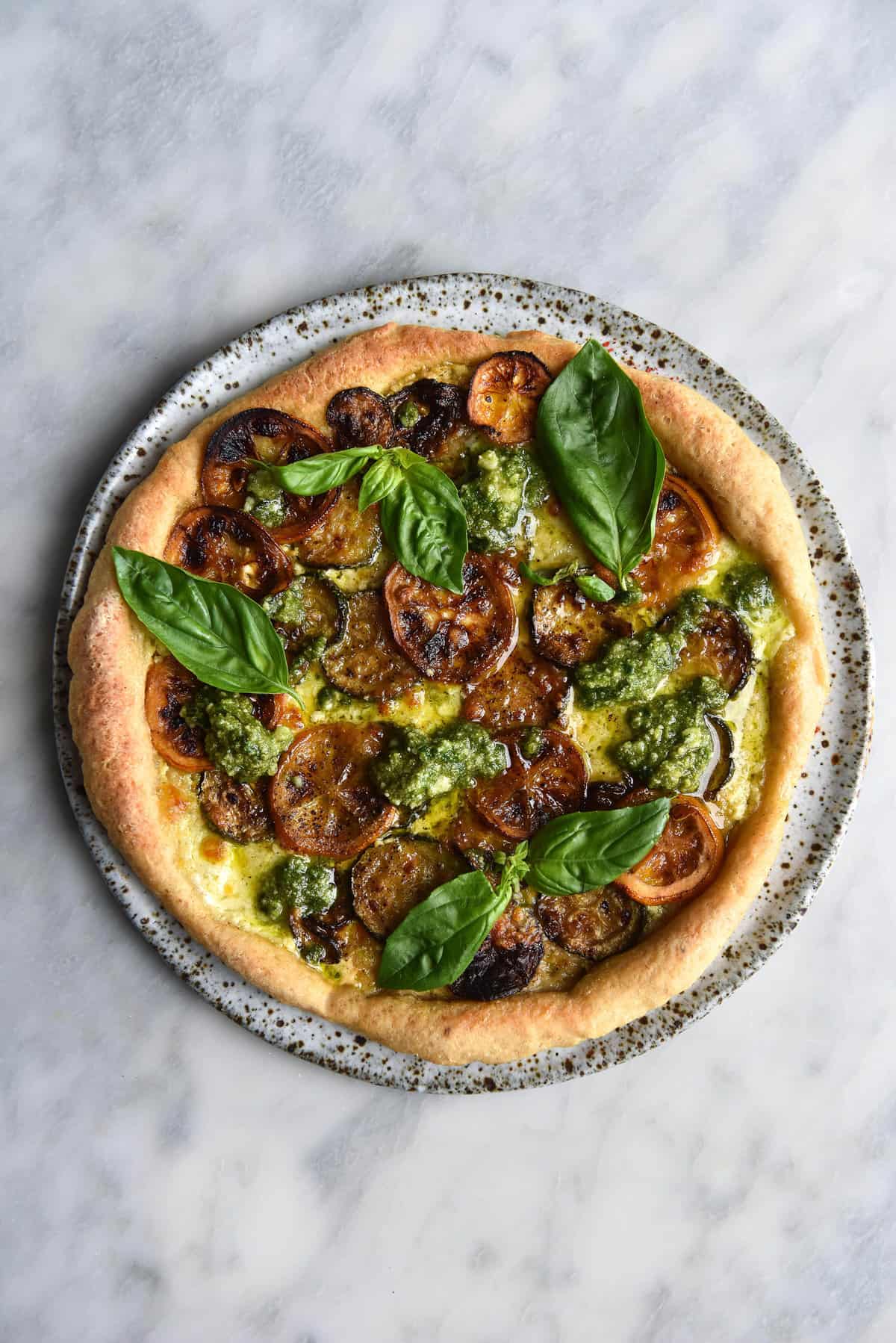 An aerial view of a gluten free pizza atop a white speckled ceramic plate on a white marble table. The pizza is topped with white sauce, basil, pesto, grilled zucchini and grilled lemon slices.